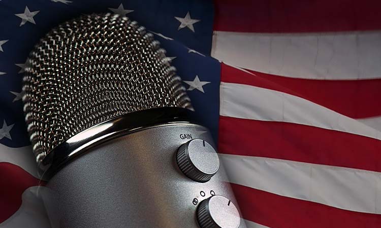 A silver microphone sits in the foreground, while a United States flag waves in the background.