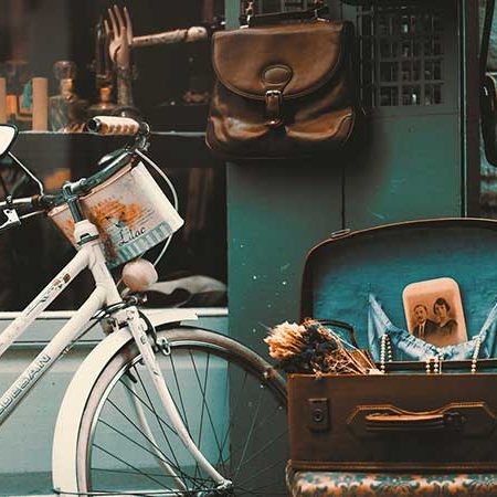 The front of an old corner store. The window displays various trinkets. A bicycle is parked outside. An open suitcase willed with nick-knacks is set on a stool. A few leather bags hang from the wall.